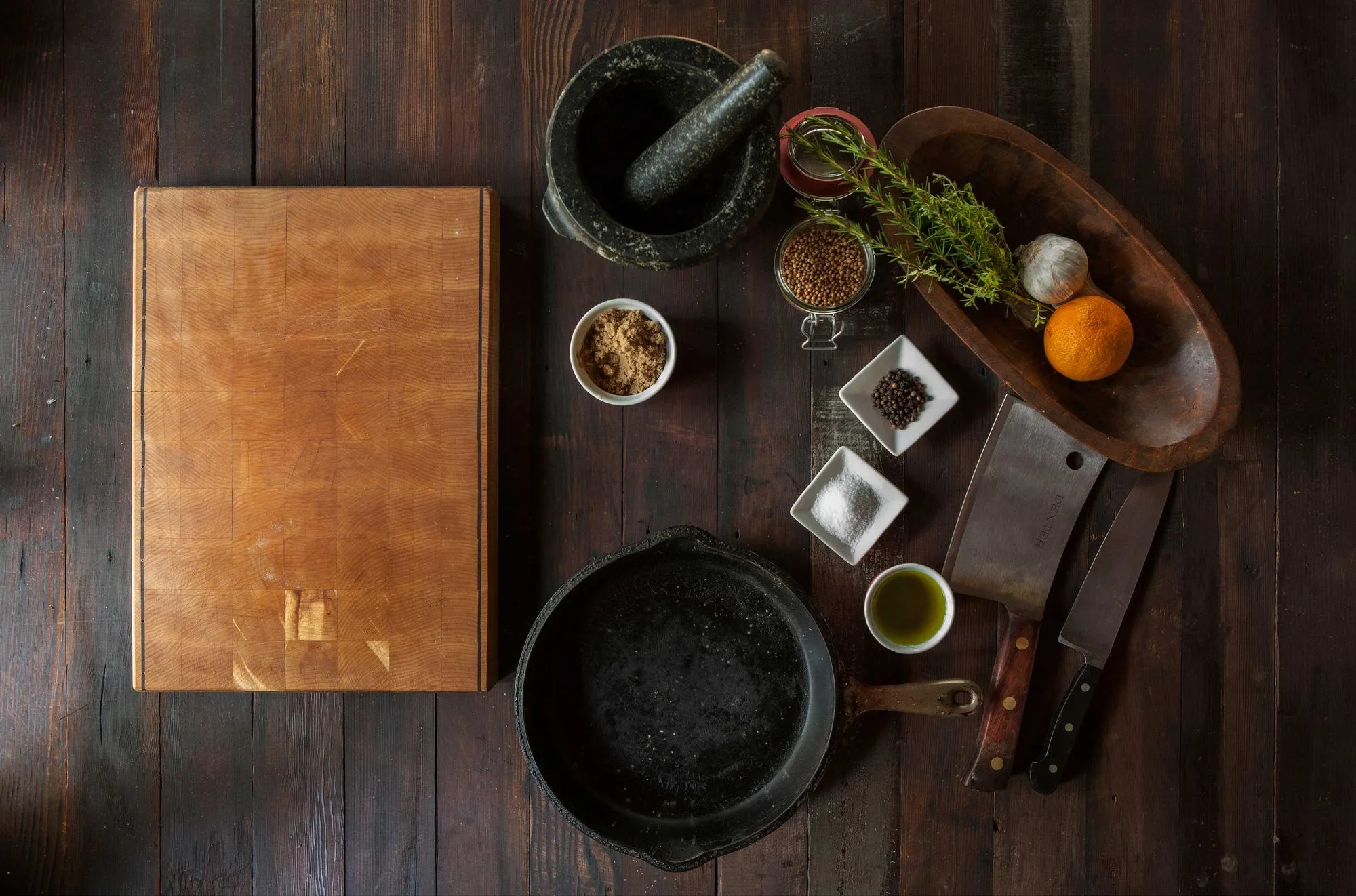 A cast iron skillet, a cleaver, a cutting board and small bowls of spices on a dark wooden table