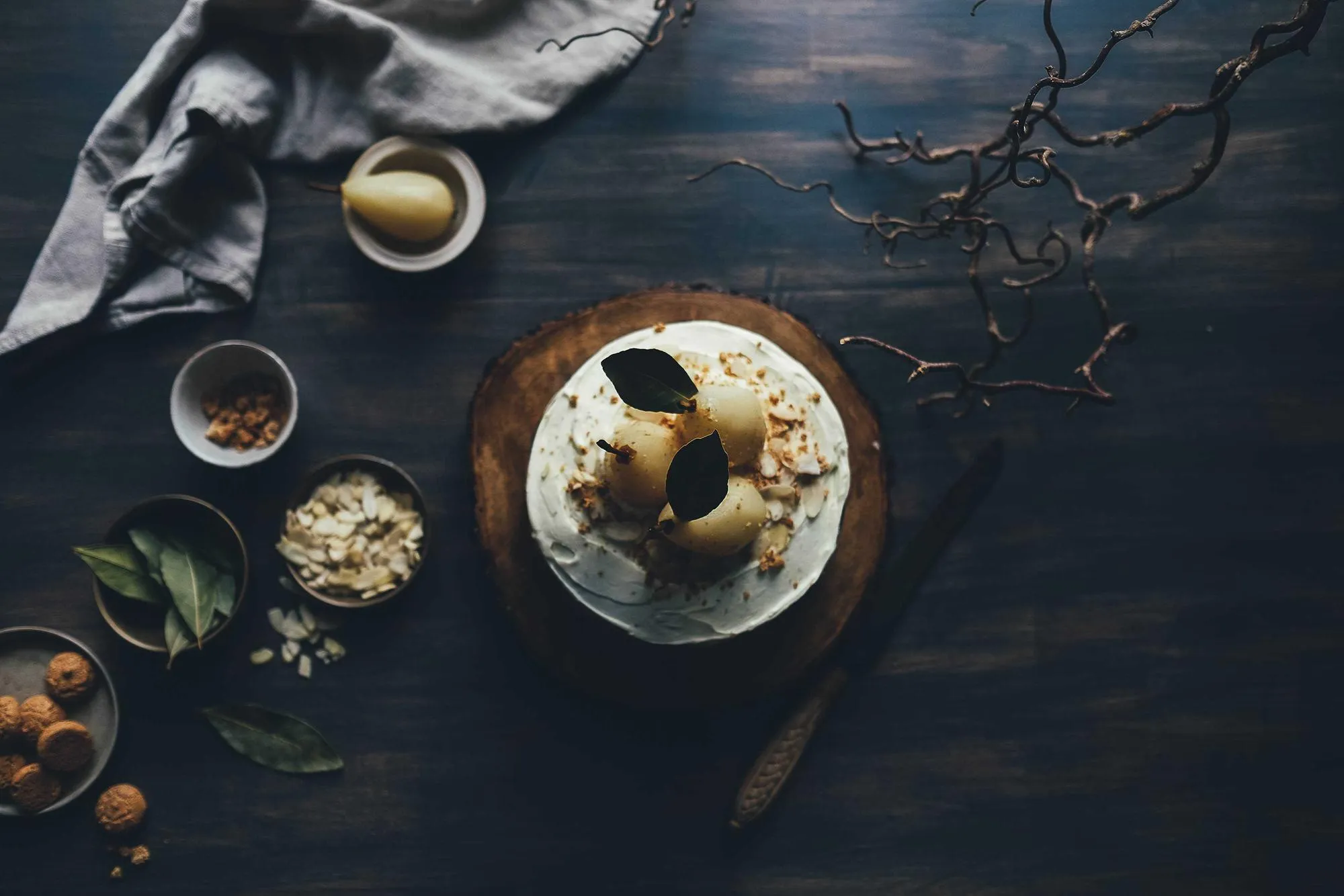 A round cake topped with roasted pears on a dark table, next to small bowls of nuts and spices