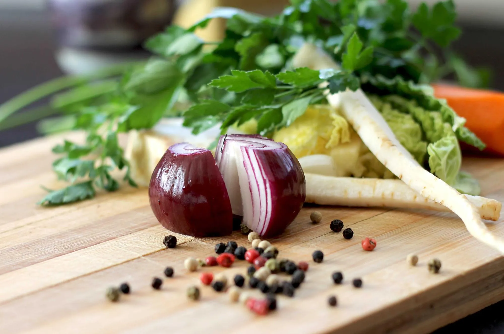 A halved red onion, parsnips, herbs and peppercorns on a wooden cutting board