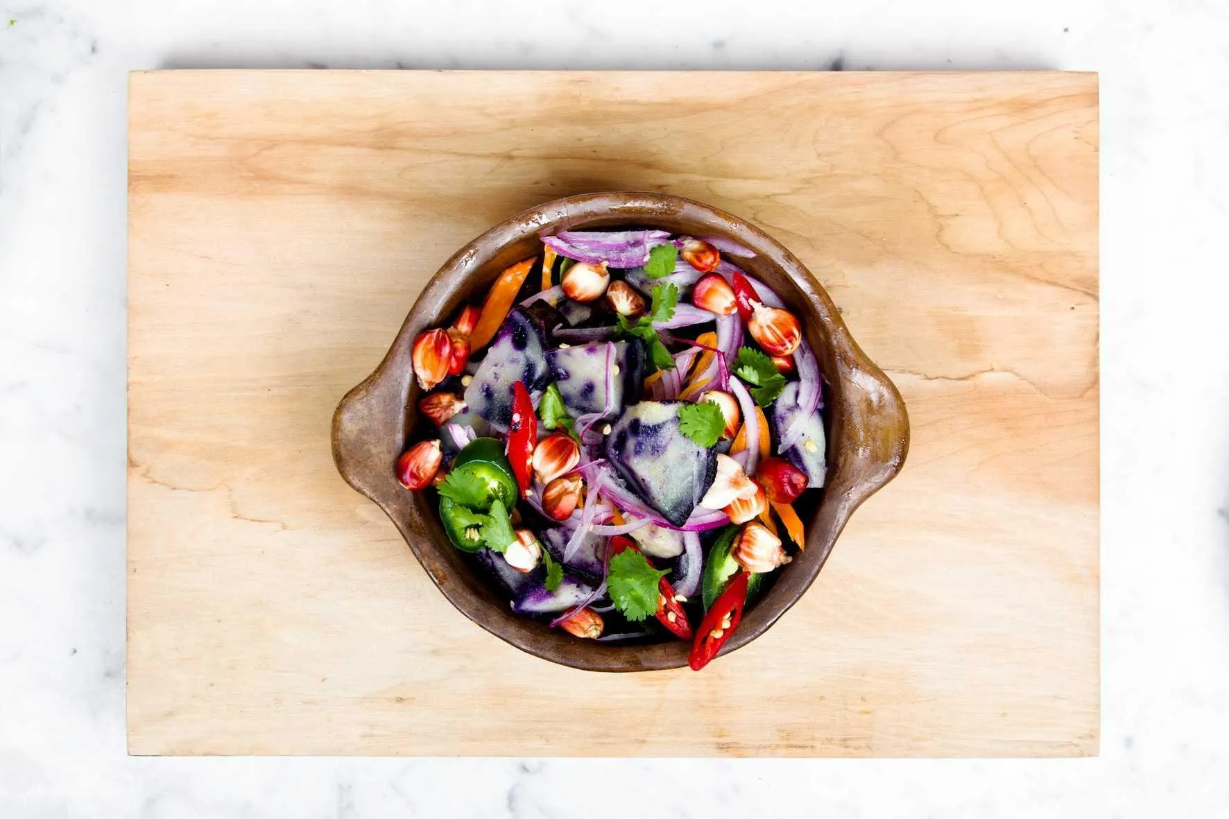 A clay bowl of sliced vegetables and herbs on a wooden board